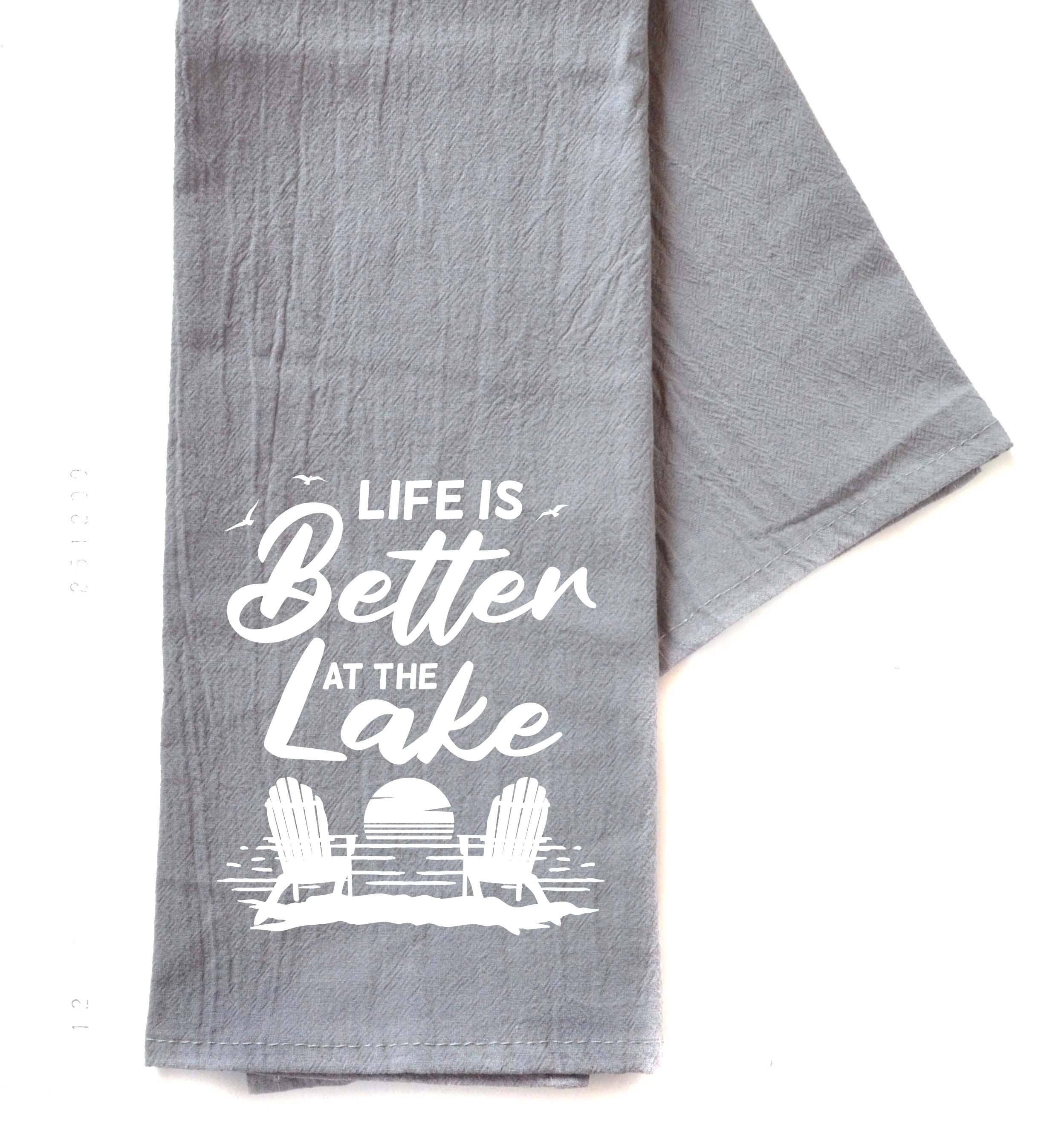 A gray cotton hand towel with the text 'LIFE IS BETTER AT THE LAKE' printed in white, along with a graphic of a chair and a lake.