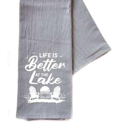 A gray cotton hand towel with the text 'LIFE IS BETTER AT THE LAKE' printed in white, along with a graphic of a chair and a lake.