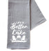 A gray cotton hand towel with the text 'LIFE IS BETTER AT THE LAKE' printed in white, along with a graphic of a chair and a lake.