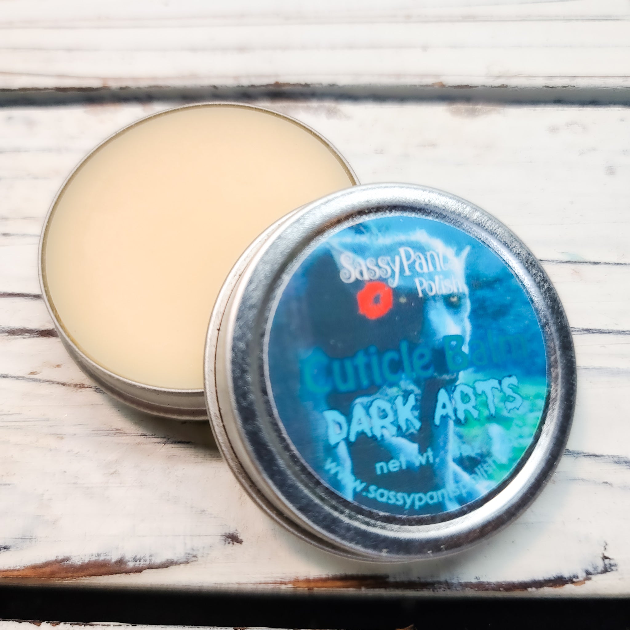 A cuticle balm open with a metal lid, showing a solid yellow cuticle balm inside, placed on a wooden surface.  Dark Arts