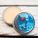 A cuticle balm open with a metal lid, showing a solid yellow cuticle balm inside, placed on a wooden surface.  Dark Arts