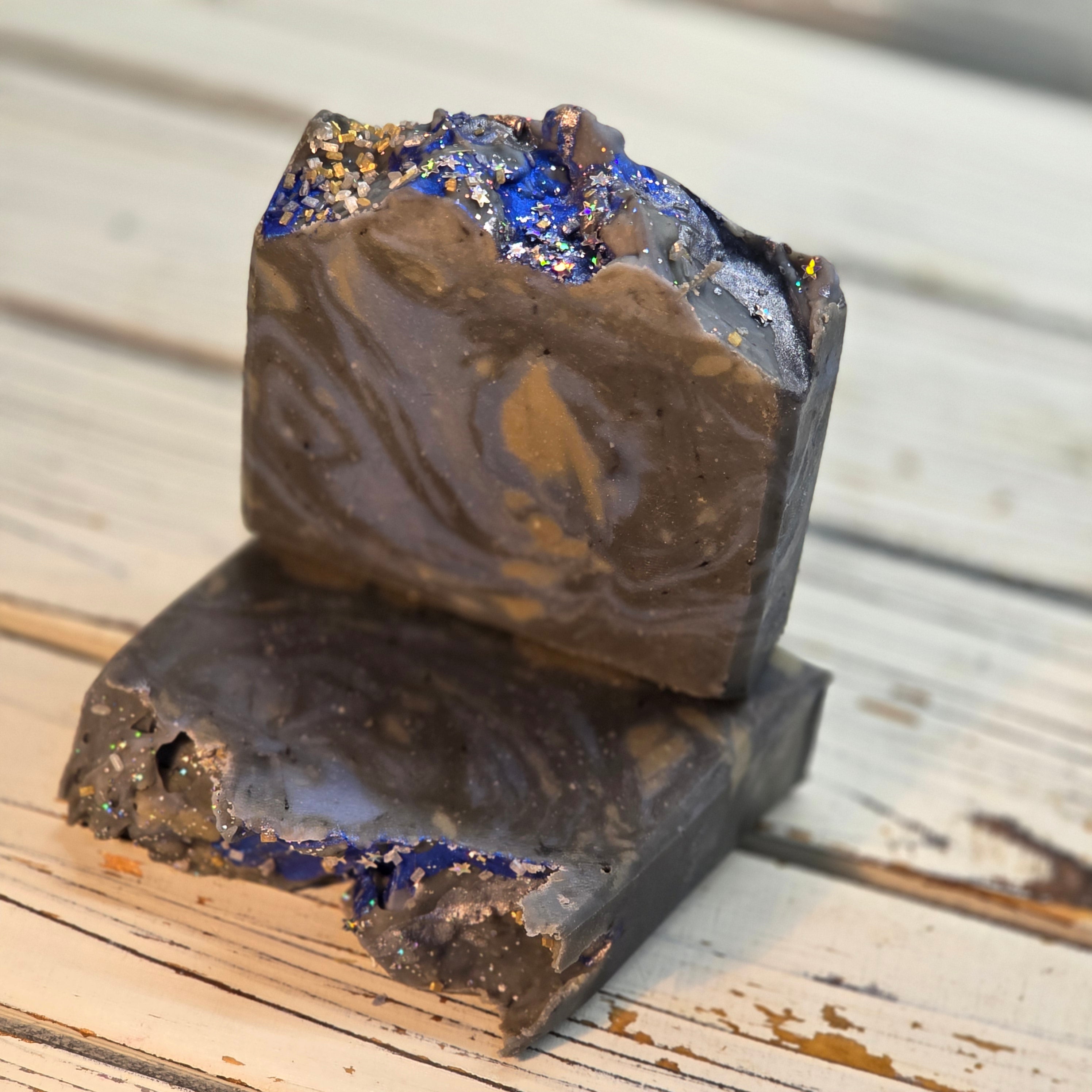 Two rough-cut dark bars of goat milk soap stacked on a whitewashed wooden surface, with visible blue glitter and metallic flecks on the exposed ends.
