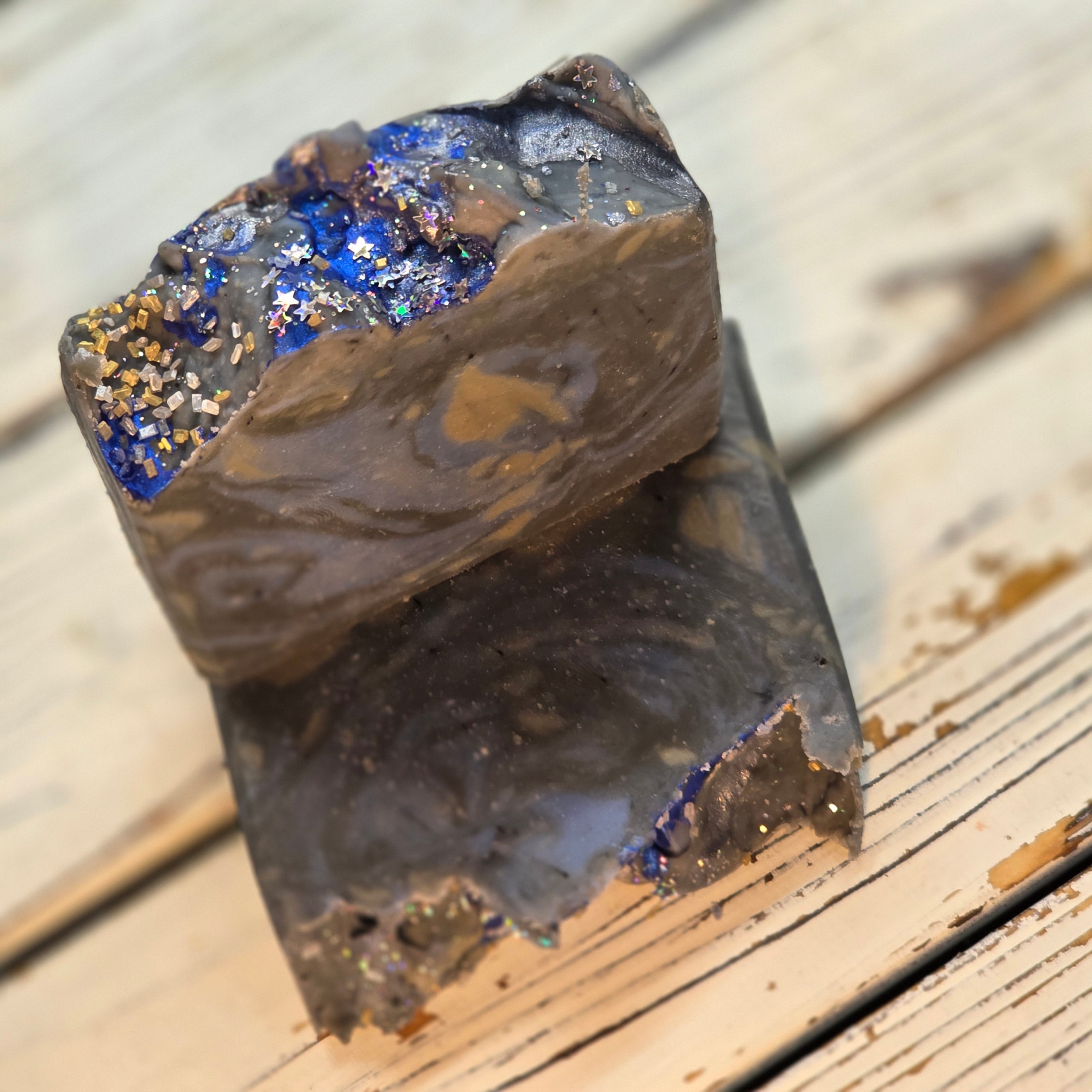 Two rustic handmade soap bars stacked on a light wood surface. The soap is dark brown with a glossy finish and features a decorative top section filled with blue glitter and gold-colored flakes.
