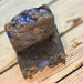 Two rustic handmade soap bars stacked on a light wood surface. The soap is dark brown with a glossy finish and features a decorative top section filled with blue glitter and gold-colored flakes.