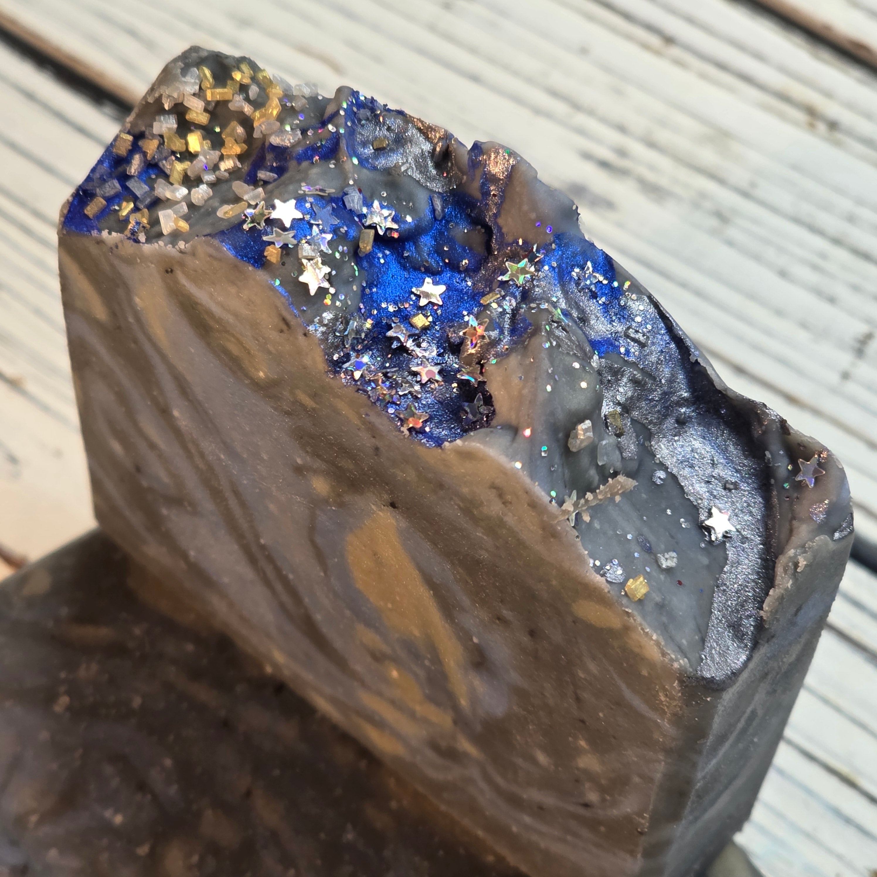 A close-up of a handmade soap bar with a rough, raw-edge top featuring a deep blue glittery interior filled with silver and gold star-shaped glitter, set against a light gray wooden background.
