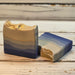 Two handmade rectangular soap bars with a marbled blue, gray, and beige design and gold leaf accents, displayed on a rustic white wooden surface.