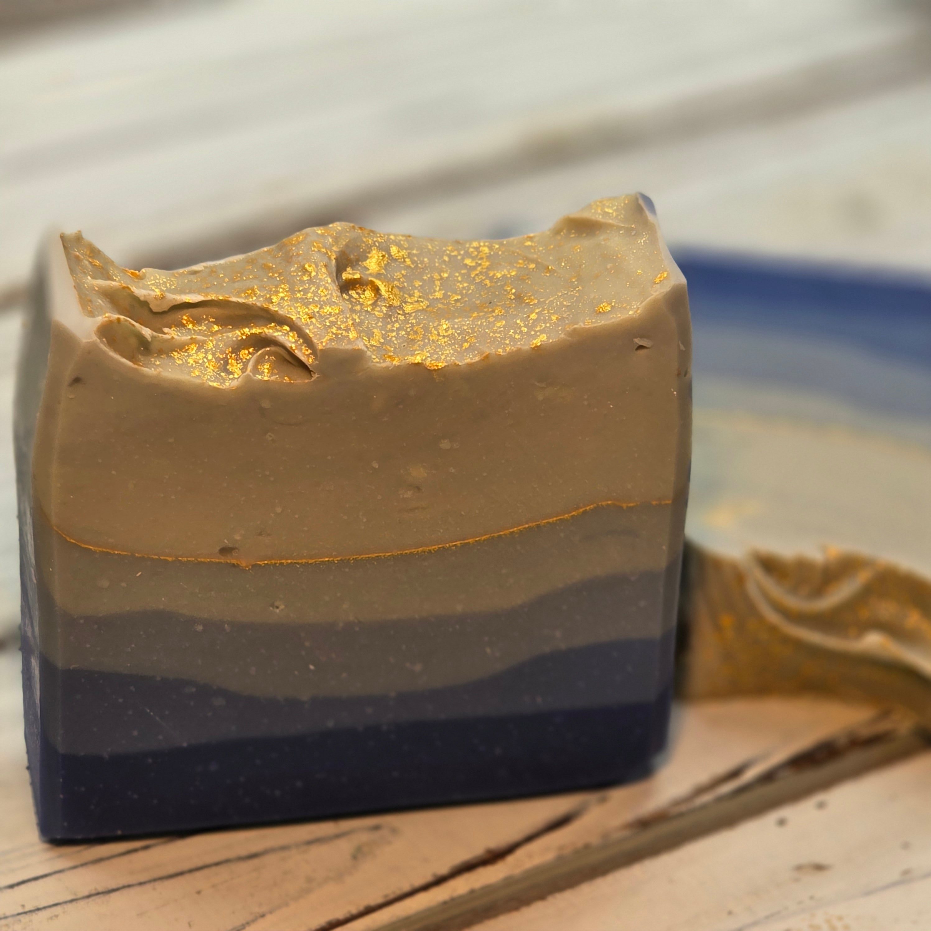Two handmade rectangular soap bars with a marbled blue, gray, and beige design and gold leaf accents, displayed on a rustic white wooden surface.