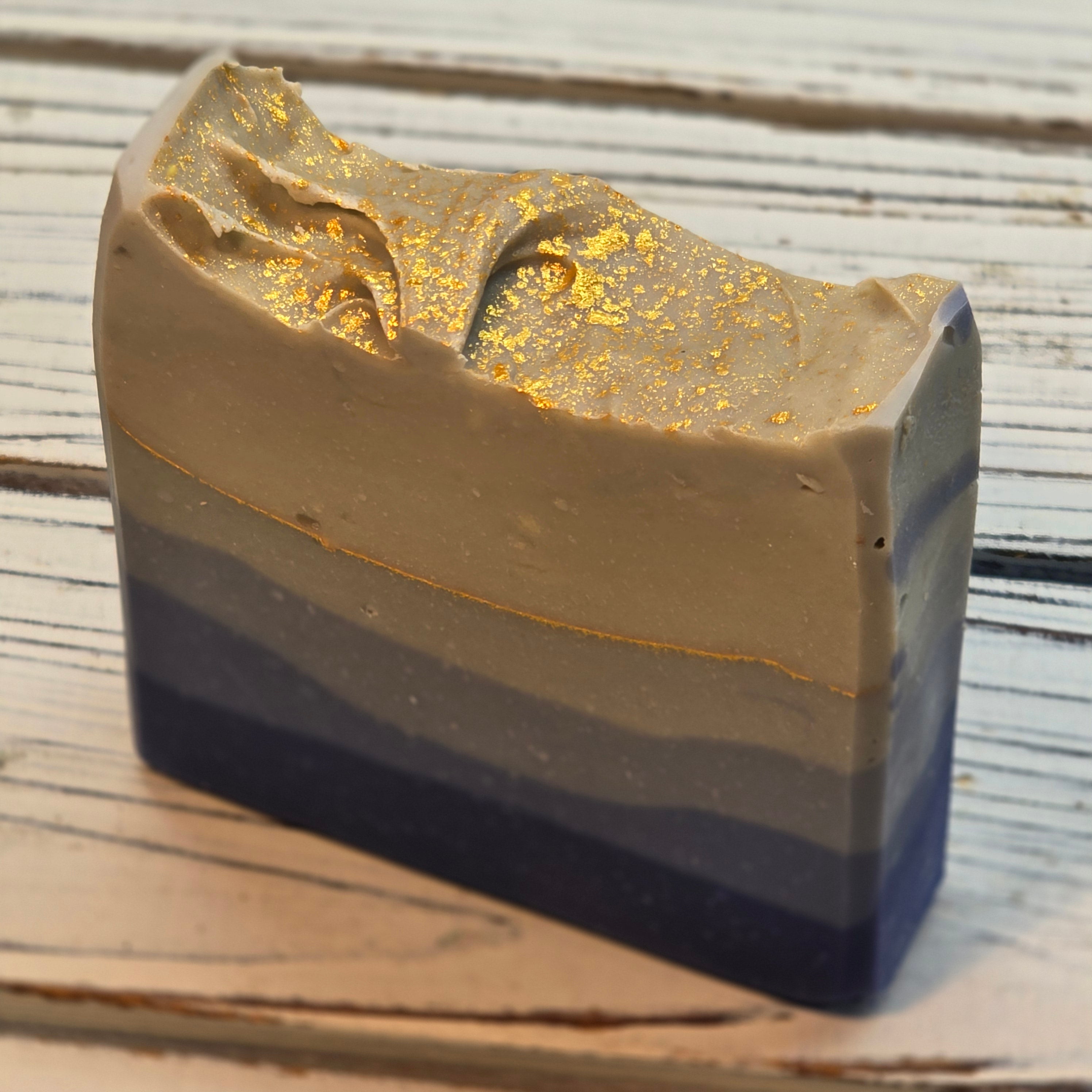 handmade rectangular soap bar with a marbled blue, gray, and beige design and gold leaf accents, displayed on a rustic white wooden surface.