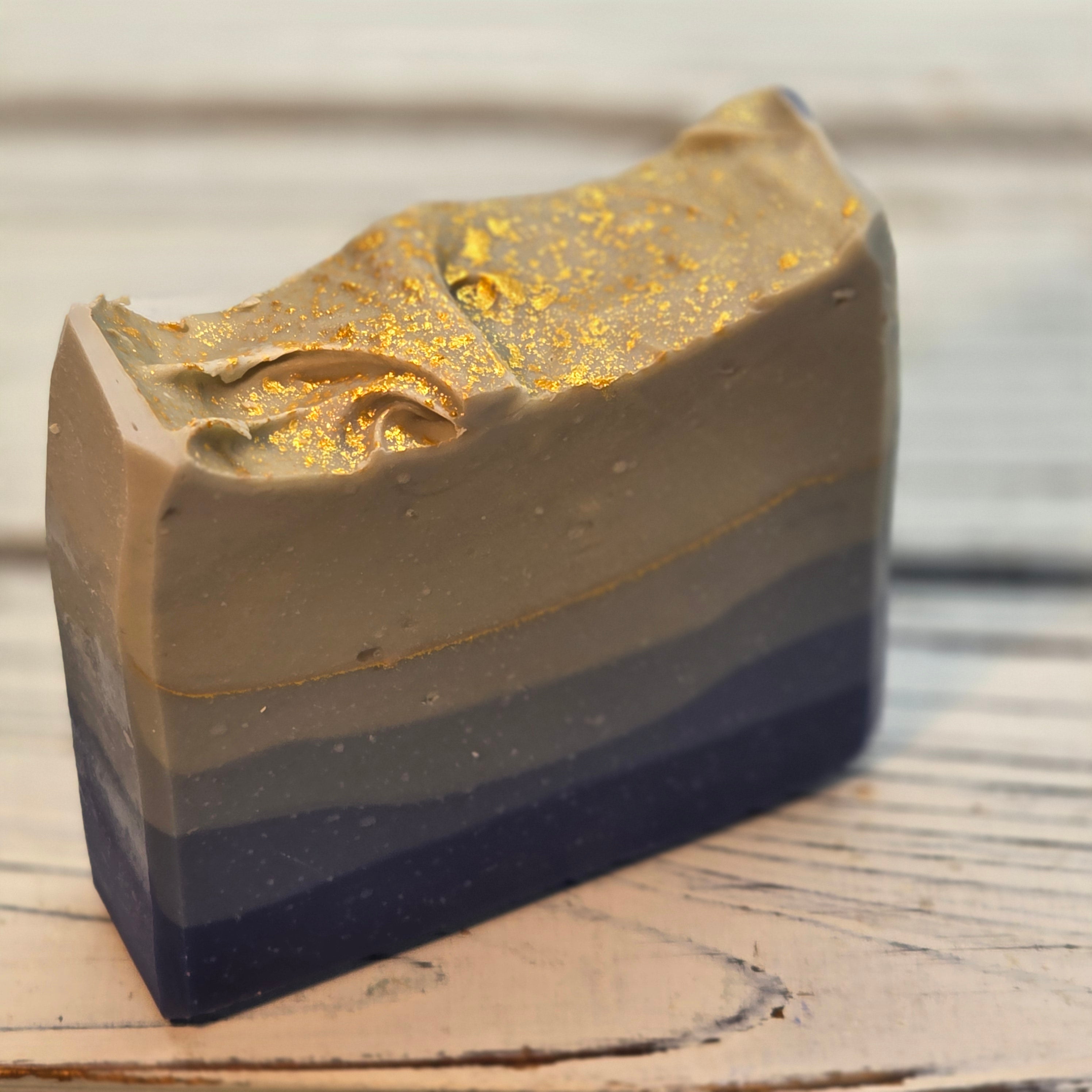 A handmade layered soap bar with a rustic, hand-drawn top edge, featuring horizontal stripes of dark navy, charcoal gray, and warm taupe, finished with a textured gold foil top.