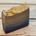 A handmade layered soap bar with a rustic, hand-drawn top edge, featuring horizontal stripes of dark navy, charcoal gray, and warm taupe, finished with a textured gold foil top.