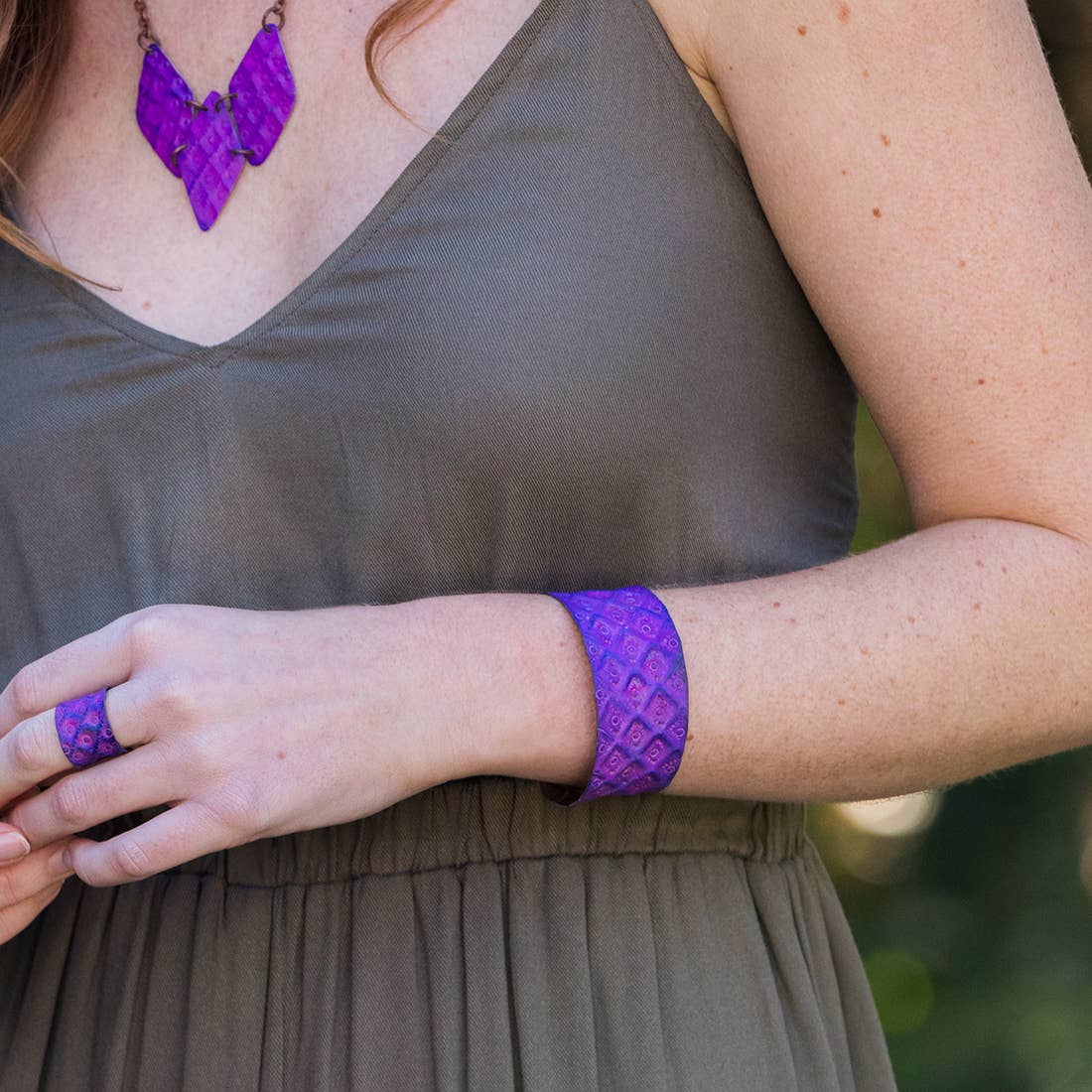 Person wearing a purple textured necklace, bracelet, and ring against a blurred green background