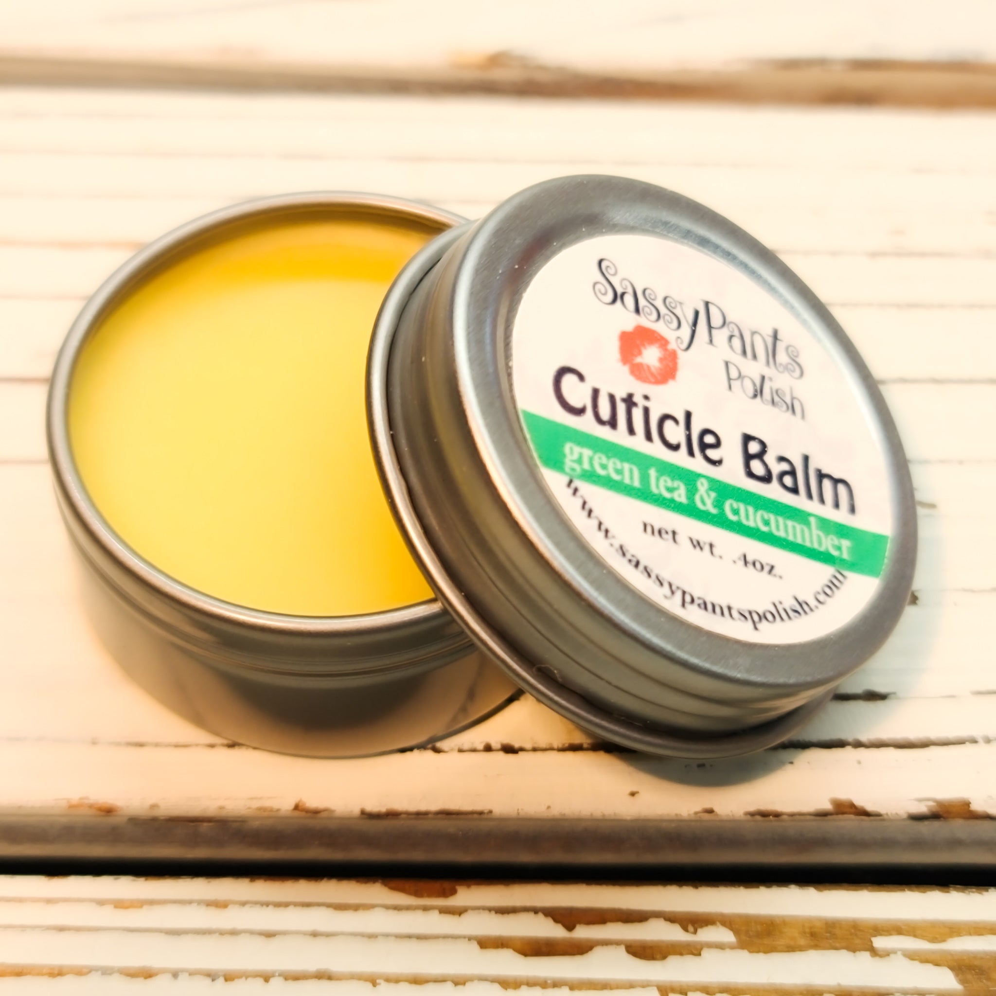 A cuticle balm open with a metal lid, showing a solid yellow cuticle balm inside, placed on a wooden surface.  Green Tea & Cucumber