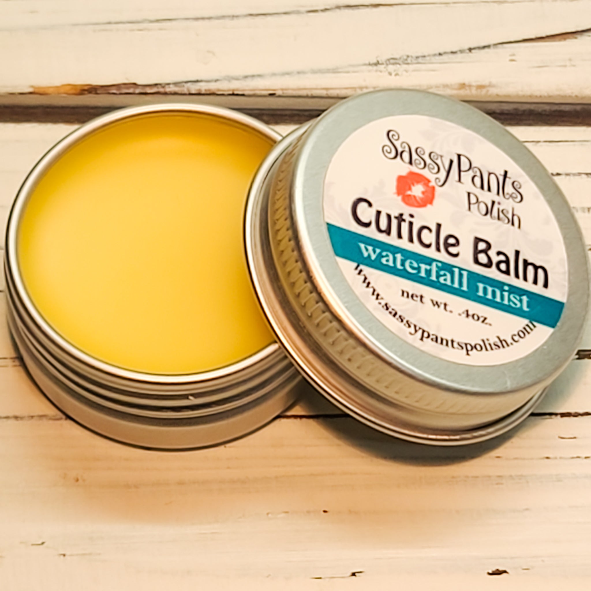 A cuticle balm open with a metal lid, showing a solid yellow cuticle balm inside, placed on a wooden surface. Waterfall Mist