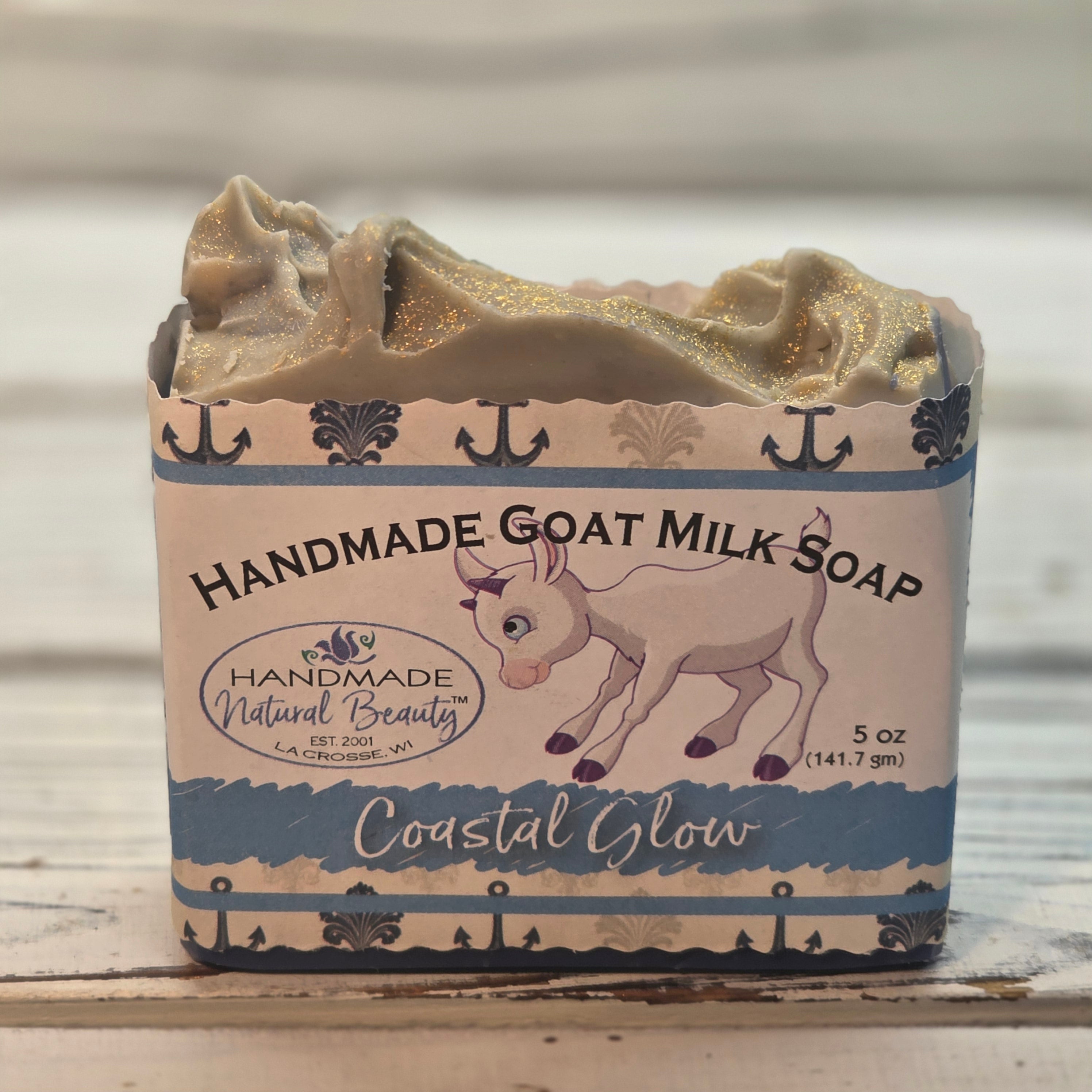 A 5 oz bar of handmade goat milk soap with a decorative paper label, resting on a rustic white wooden surface. The soap has a textured, glittery beige-gold finish, and the label features an illustration of a goat and the brand name 'Handmade Natural Beauty'.