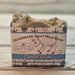 A 5 oz bar of handmade goat milk soap with a decorative paper label, resting on a rustic white wooden surface. The soap has a textured, glittery beige-gold finish, and the label features an illustration of a goat and the brand name 'Handmade Natural Beauty'.