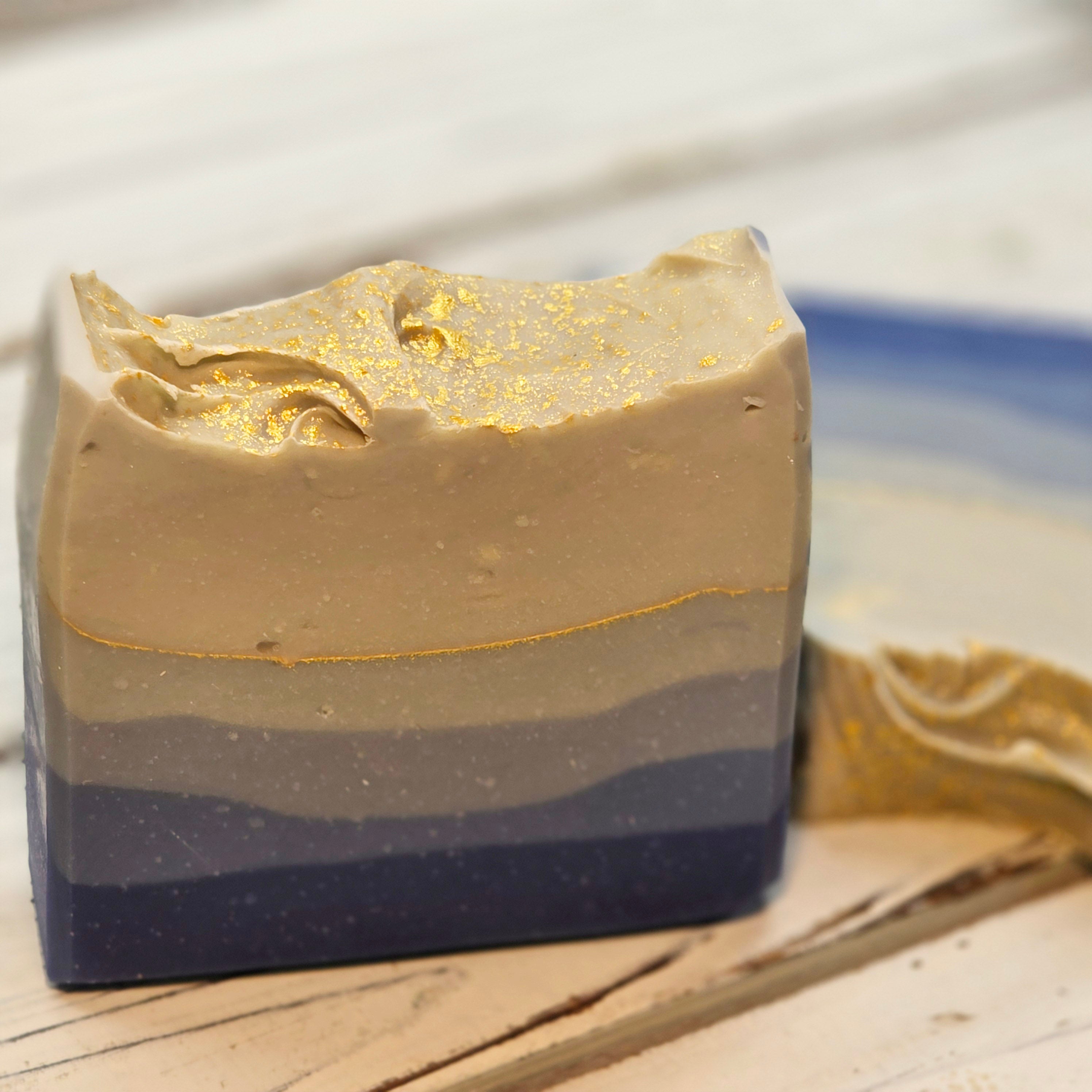 A handmade ombre blue soap bar with a gold glitter finish on top, resting on a light-colored wooden surface.