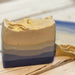 A handmade ombre blue soap bar with a gold glitter finish on top, resting on a light-colored wooden surface.