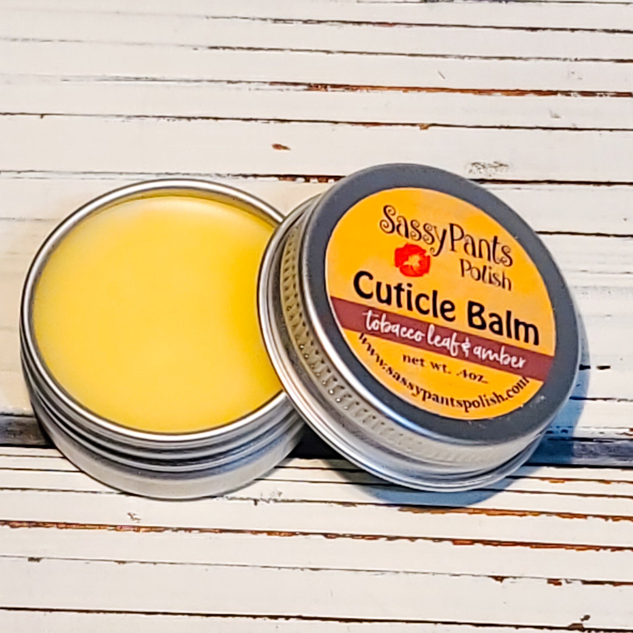 A cuticle balm open with a metal lid, showing a solid yellow cuticle balm inside, placed on a wooden surface.  Tobacco Leaf & Amber by Handmade Natural Beauty