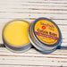 A cuticle balm open with a metal lid, showing a solid yellow cuticle balm inside, placed on a wooden surface.  Tobacco Leaf & Amber by Handmade Natural Beauty