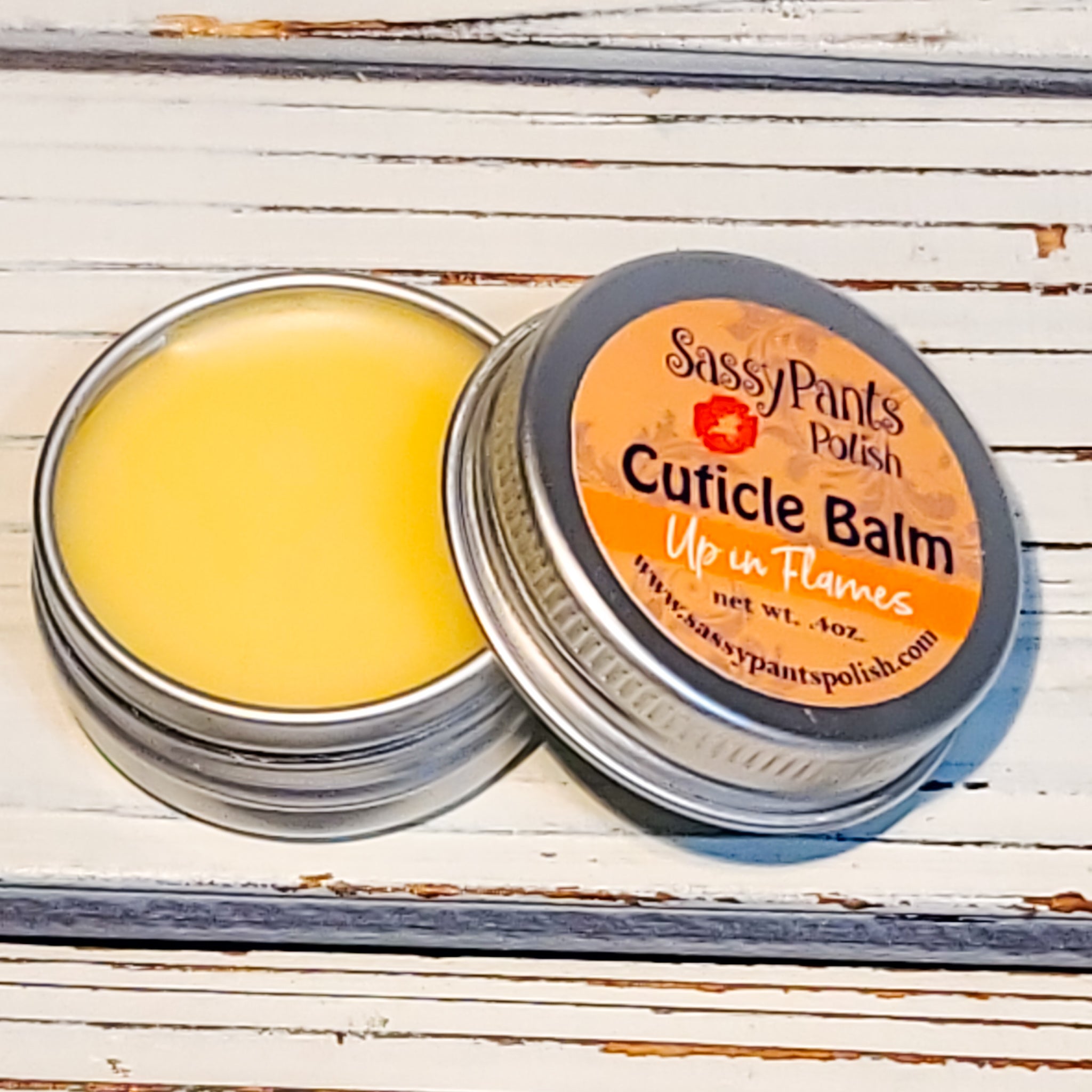A cuticle balm open with a metal lid, showing a solid yellow cuticle balm inside, placed on a wooden surface.  Up in Flames by Sassy Pants Polish