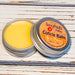 A cuticle balm open with a metal lid, showing a solid yellow cuticle balm inside, placed on a wooden surface.  Up in Flames by Sassy Pants Polish