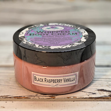 A jar of shea butter whipped body butter labeled 'Black Raspberry Vanilla' by Handmade Natural Beauty., featuring a dusty rose-brown product inside a black container, set against a rustic white wooden background.