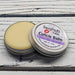 A cuticle balm open with a metal lid, showing a solid yellow cuticle balm inside, placed on a wooden surface. Stress Less Lavender