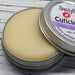 A cuticle balm open with a metal lid, showing a solid yellow cuticle balm inside, placed on a wooden surface. Stress Less Lavender.
