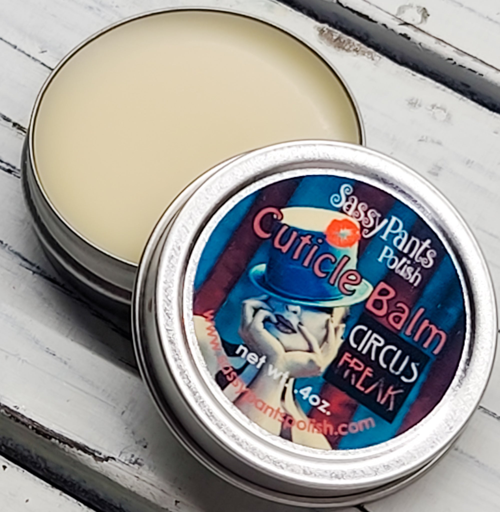 A cuticle balm open with a metal lid, showing a solid yellow cuticle balm inside, placed on a wooden surface. Circus Freak