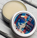 A cuticle balm open with a metal lid, showing a solid yellow cuticle balm inside, placed on a wooden surface. Circus Freak