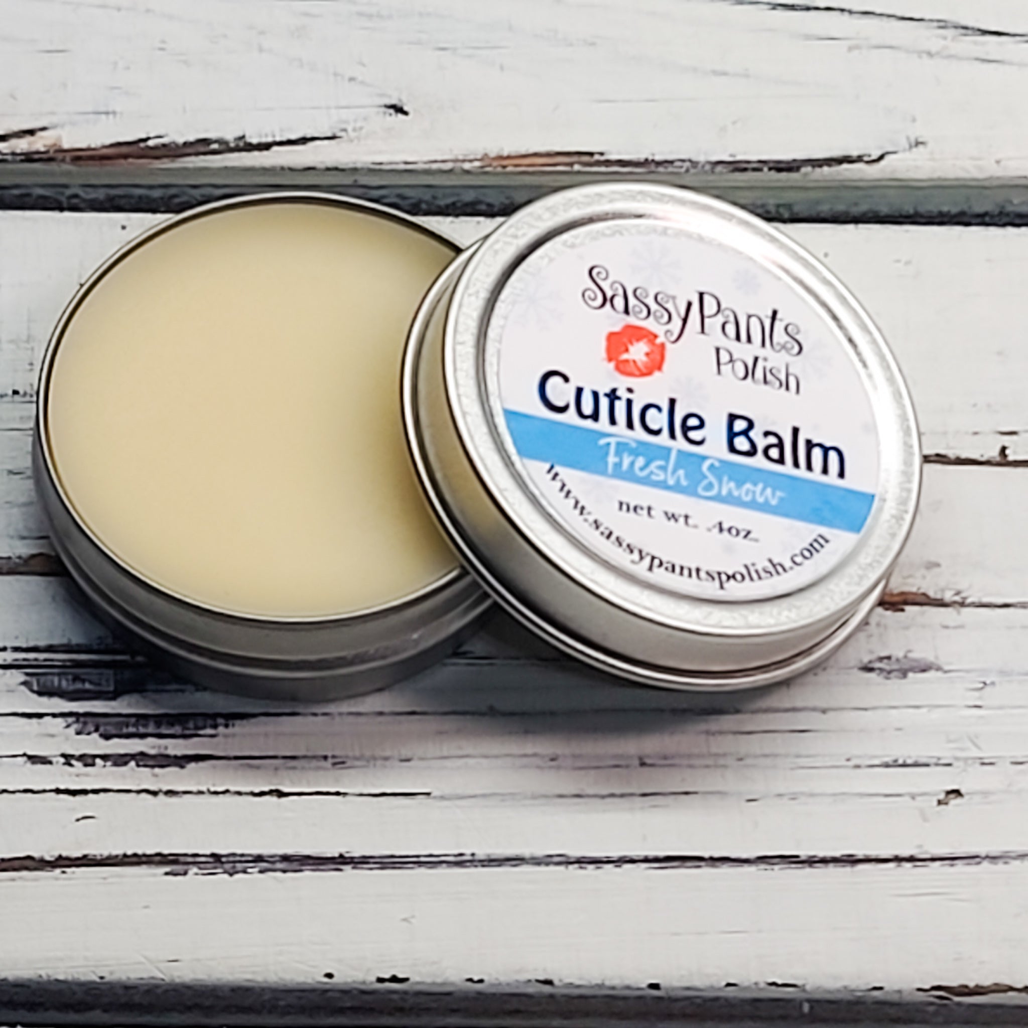 A cuticle balm open with a metal lid, showing a solid yellow cuticle balm inside, placed on a wooden surface.  Fresh Snow