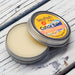 A cuticle balm open with a metal lid, showing a solid yellow cuticle balm inside, placed on a wooden surface.  Fruity Loops
