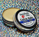 A cuticle balm open with a metal lid, showing a solid yellow cuticle balm inside, placed on a wooden surface. Arabian Nights